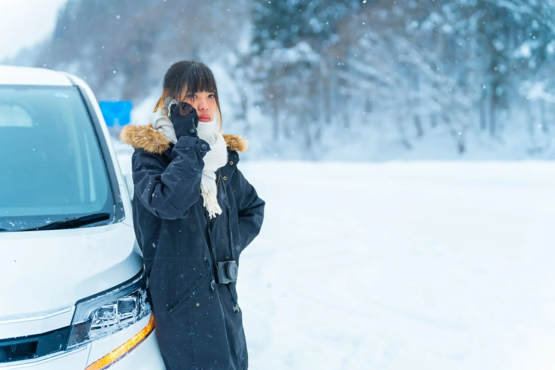femme appuyée sur sa voiture sur une route enneigée qui tente d'appeler une assistance