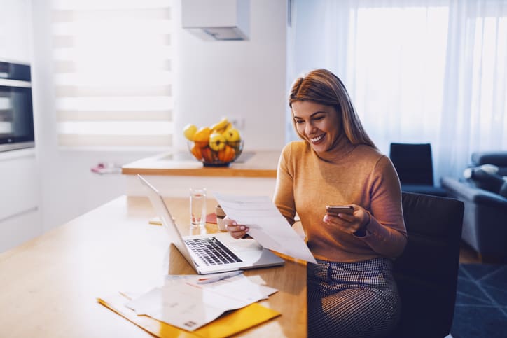 Femme blonde souriante qui regarde un document et dans l'autre mains se trouve son téléphone. Sur la table sont ordinateur portable est posé.