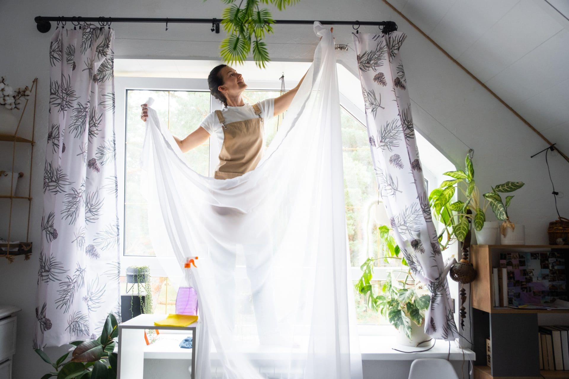 La femme en tablier accroche des rideaux de tulle transparent sur de grandes fenêtres mansardées de la maison à l’intérieur de l’intérieur